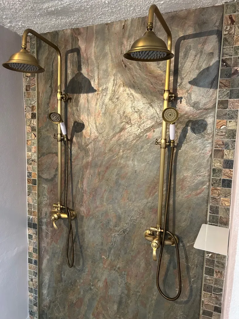 Deux colonnes de douche dorées côte à côte, devant un mur de pierre encadré de mosaïque